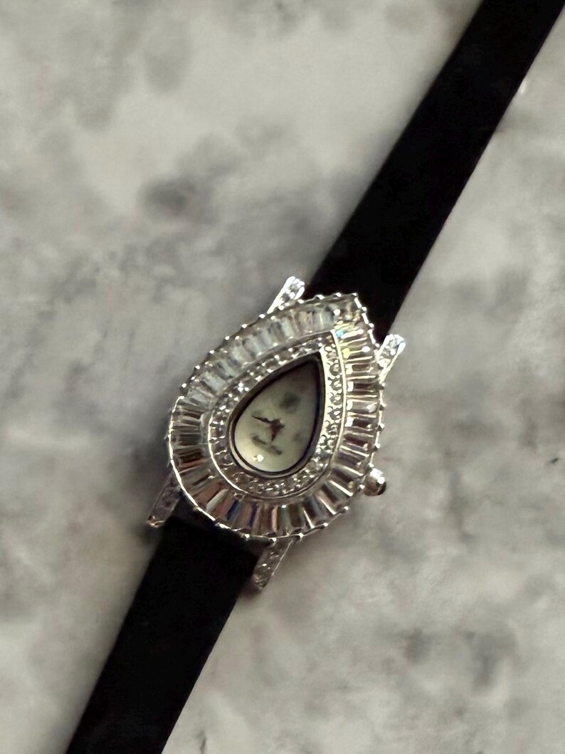May include: A silver-toned wristwatch featuring a teardrop-shaped case surrounded by a border of baguette-cut clear crystals. The watch face is white with thin hands and is attached to a slim, solid black strap that extends vertically.