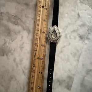 May include: A black watch with a teardrop-shaped face surrounded by clear crystals, featuring a black strap with a crystal-encrusted buckle. A wooden ruler placed alongside shows the watch measures approximately 8.5 inches in total length.