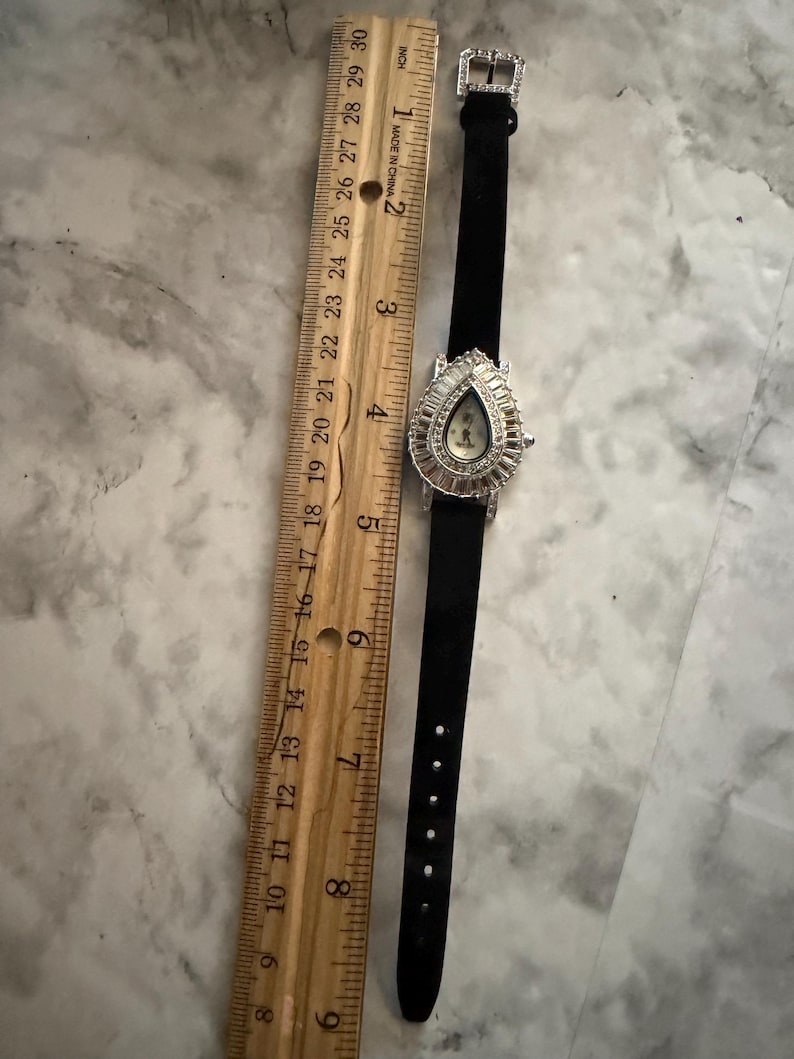 May include: A black watch with a teardrop-shaped face surrounded by clear crystals, featuring a black strap with a crystal-encrusted buckle. A wooden ruler placed alongside shows the watch measures approximately 8.5 inches in total length.