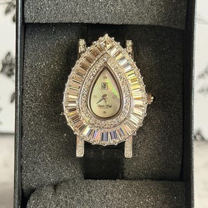 May include: A teardrop-shaped wristwatch featuring a silver-tone case encrusted with baguette and round clear crystals. The dial is mother-of-pearl with the logo W and the text Pierre Wick Beverly Hills. It includes a black strap and a crown.