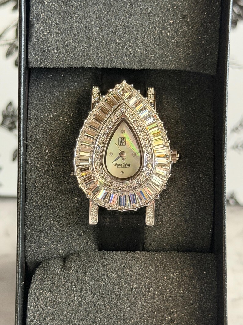 May include: A teardrop-shaped wristwatch featuring a silver-tone case encrusted with baguette and round clear crystals. The dial is mother-of-pearl with the logo W and the text Pierre Wick Beverly Hills. It includes a black strap and a crown.