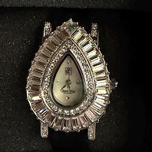 May include: A teardrop-shaped wristwatch featuring a silver-tone metal case encrusted with baguette and round-cut clear crystals. The white dial displays the Victoria Wieck Beverly Hills logo and initials, with silver hands and crystal hour markers.
