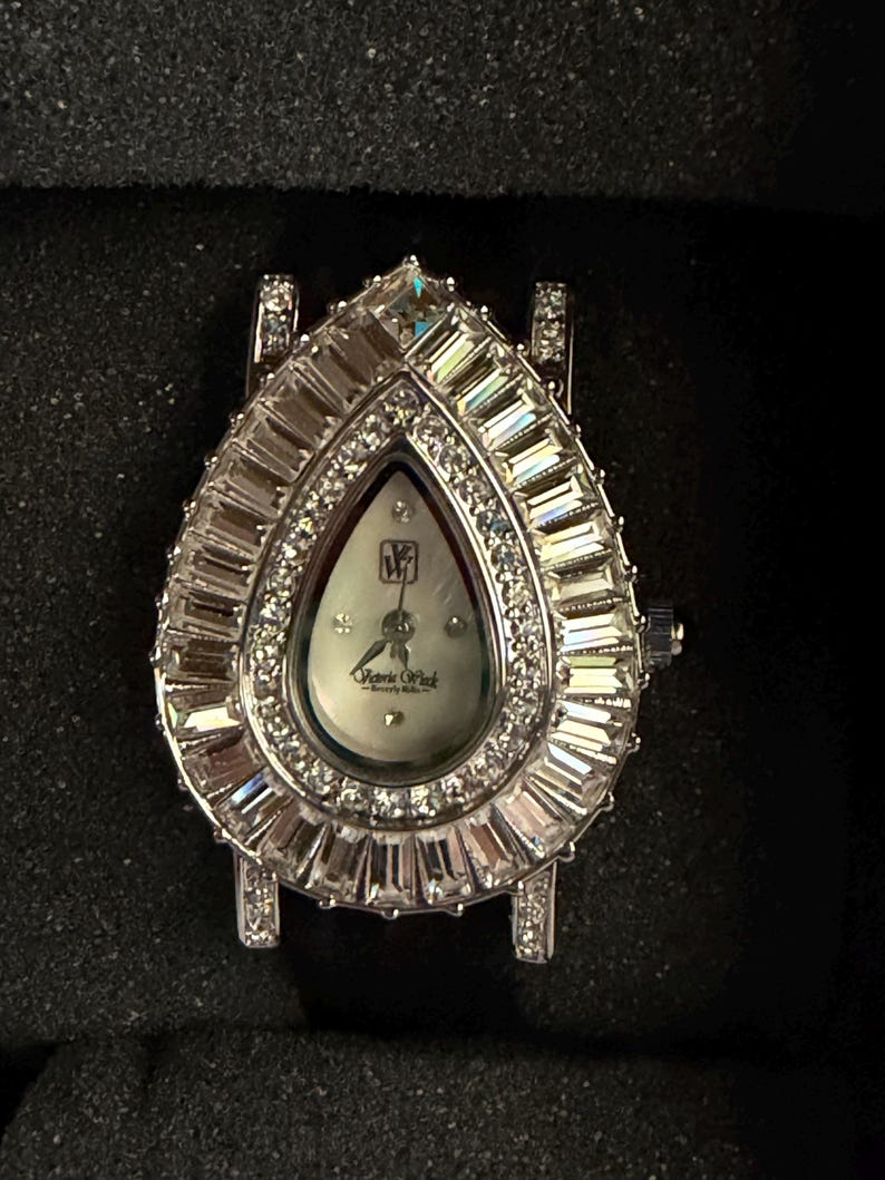 May include: A teardrop-shaped wristwatch featuring a silver-tone metal case encrusted with baguette and round-cut clear crystals. The white dial displays the Victoria Wieck Beverly Hills logo and initials, with silver hands and crystal hour markers.