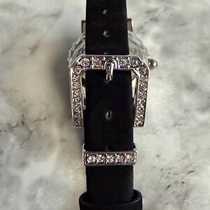 May include: A black watch strap featuring a silver-toned buckle and a matching keeper, both encrusted with small, clear, sparkling crystals. The strap is made of a dark, matte material and includes a series of adjustment holes for a secure fit.