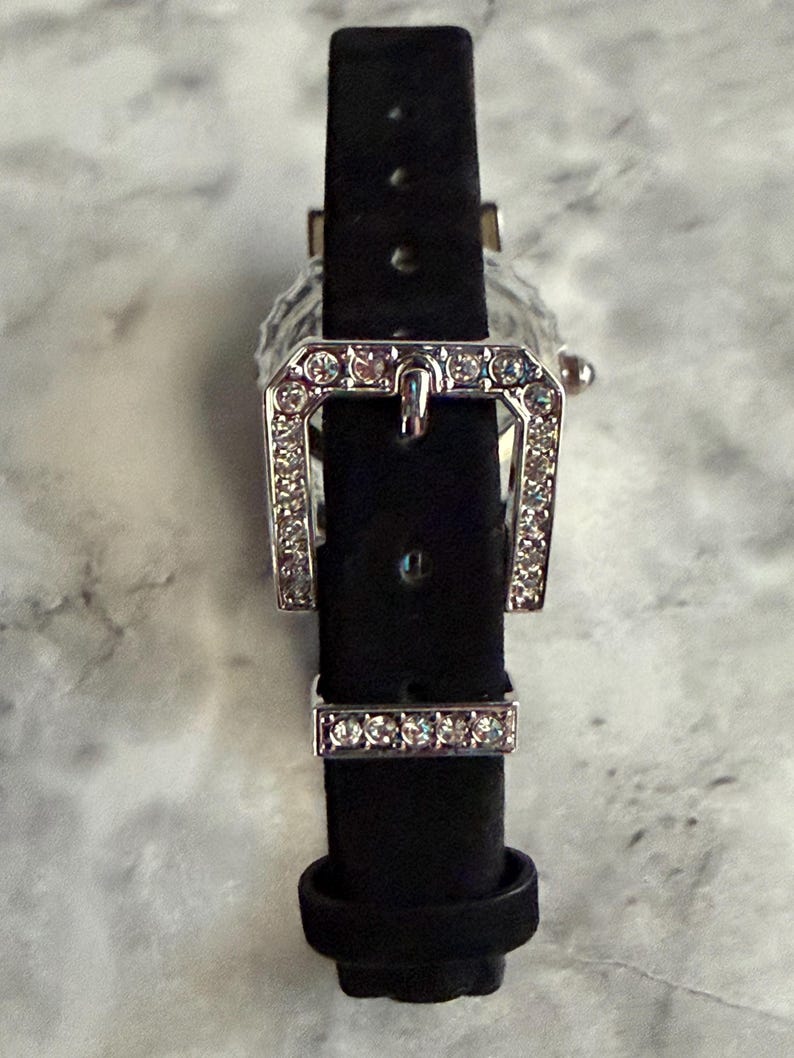 May include: A black watch strap featuring a silver-toned buckle and a matching keeper, both encrusted with small, clear, sparkling crystals. The strap is made of a dark, matte material and includes a series of adjustment holes for a secure fit.