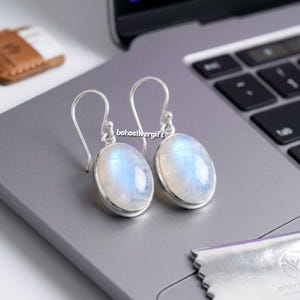 May include: A pair of silver drop earrings featuring oval-shaped moonstones with a blue iridescent sheen. The earrings have curved silver ear wires and the text bohosilvergift is visible between the two stones on the surface.