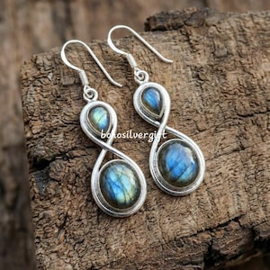 May include: A pair of silver earrings with a teardrop-shaped top and an oval bottom, each containing a blue and green iridescent stone. The earrings are displayed on a textured wooden surface. The text "bolosilvergift" is visible.
