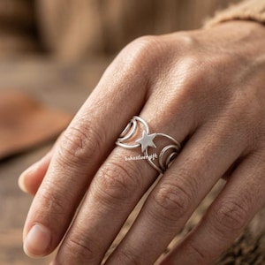 May include: A silver ring featuring a star design at the center, flanked by curved, openwork details. The ring is worn on a finger, showcasing its intricate design. The ring's band has a unique, open design. The ring is a piece of jewelry.