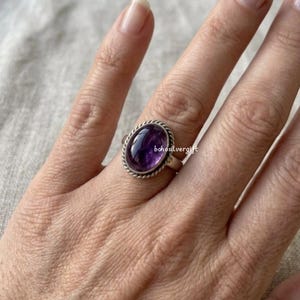 Könnte beinhalten: Ein silberner Ring mit einem ovalen, tiefvioletten Edelstein. Der Edelstein ist in einer silbernen Lünette mit einem gedrehten Seildesign eingefasst. Der Ring befindet sich am Finger einer Person.
