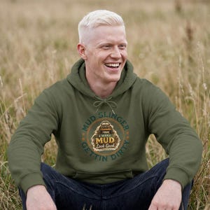 May include: An olive green hoodie with a graphic that reads "Mud Slinger Gettin' Dirty I Make Mud Look Good." The design features a truck illustration. The person is sitting in a field of tall grass.