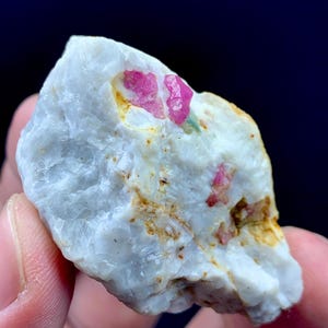 May include: A close-up of a rough, white mineral specimen with vibrant pink ruby crystals embedded within. The stone has a textured surface with hints of yellow and brown, and the ruby crystals are a striking contrast.