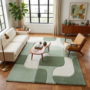 May include: A rectangular area rug featuring a geometric pattern of interlocking shapes in varying shades of sage green, olive green, and cream white. The rug has a textured, plush pile surface and is positioned on a wooden floor in a living room.