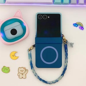May include: A blue phone case with a matching strap, a pink cat-shaped case, and several decorative stickers, including a rainbow, a moon, a frog, and a bear. A keyboard is in the background.
