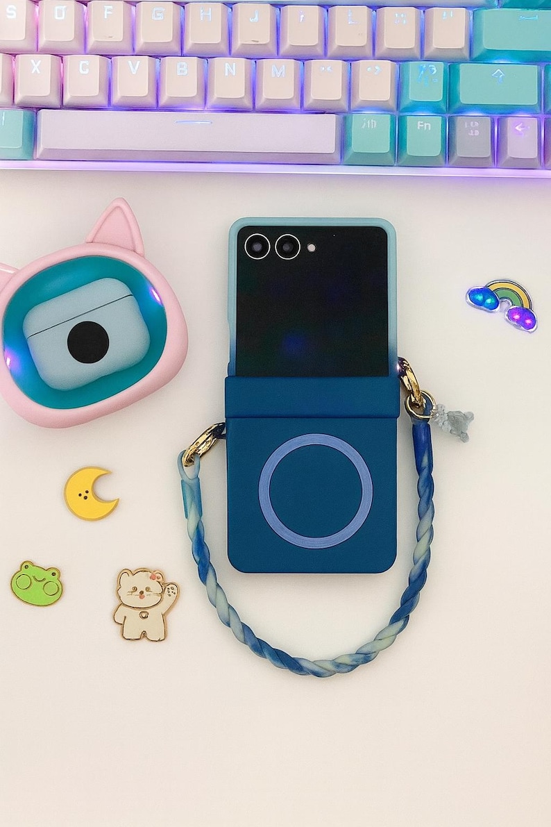 May include: A blue phone case with a matching strap, a pink cat-shaped case, and several decorative stickers, including a rainbow, a moon, a frog, and a bear. A keyboard is in the background.