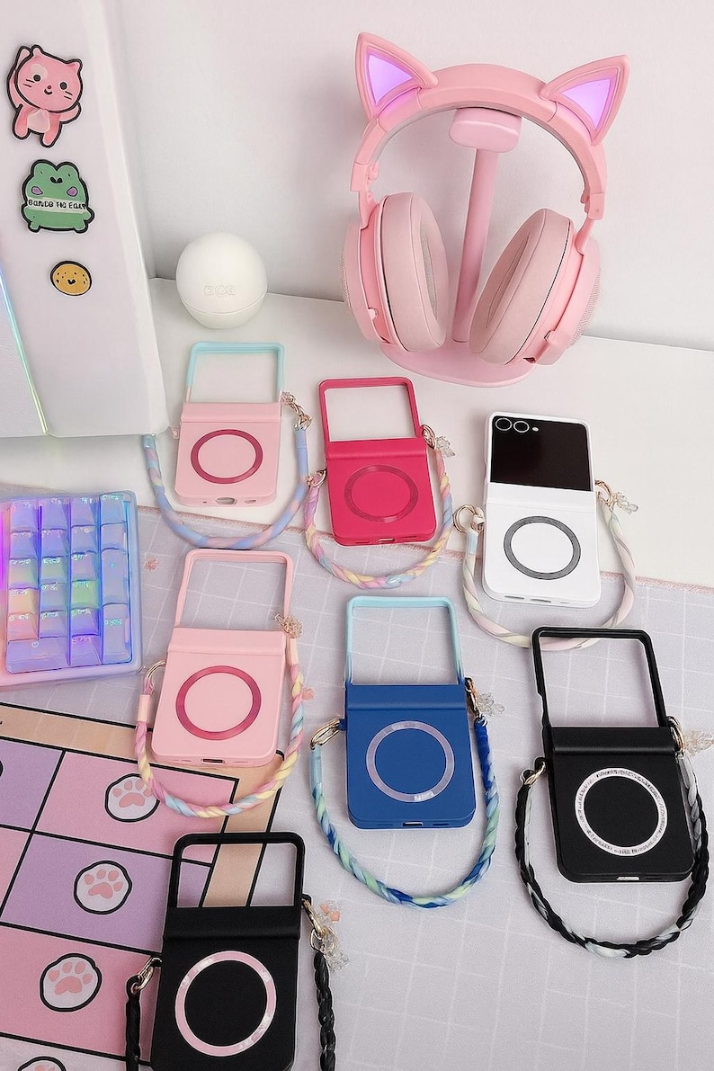 May include: A collection of phone cases in various colors, including pink, red, blue, and black, each with a circular design. A pink cat ear headset sits on a stand. A keyboard and stickers are also visible.