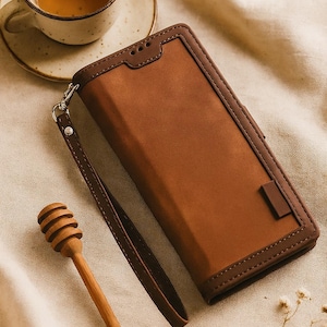 May include: A brown leather phone case with a wrist strap, featuring a darker brown trim. A wooden honey dipper and a cup of tea on a saucer are also in the image. The case has a rectangular design.