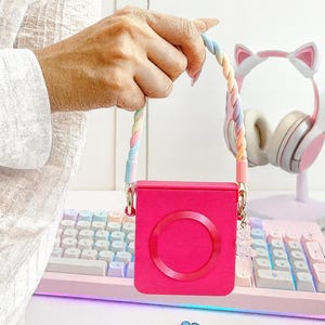 May include: A bright pink square case with a circular design, held by a rainbow-colored braided strap. The case has gold-colored hardware and a small charm. A pink and white keyboard and cat-ear headphones are in the background.