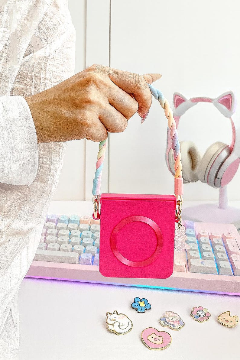 May include: A bright pink square case with a circular design, held by a rainbow-colored braided strap. The case has gold-colored hardware and a small charm. A pink and white keyboard and cat-ear headphones are in the background.