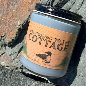 Peut inclure: Une bougie en cire de soja grise dans un pot en verre avec un couvercle en métal noir. L'étiquette présente un fond imitation bois, une illustration de plongeon noir, des pins verts et le texte I'M COMING TO THE COTTAGE, Cardamom & Tobacco.
