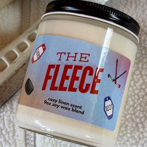 Peut inclure: Une bougie en pot de verre avec un couvercle noir et une étiquette bleu clair portant le texte THE FLEECE, cozy linen scent, 9oz soy wax blend, ainsi que des illustrations de crosses de hockey, d'un palet et deux étiquettes numérotées.