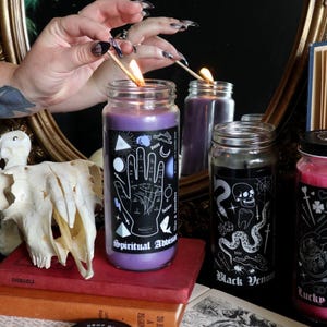 Peut inclure: Une collection de bougies en pot de verre avec des étiquettes noires et des illustrations graphiques blanches. Les étiquettes portent des textes comme Spiritual Advisor, Black Venom et Lucky in Love. Les bougies contiennent des cires de couleurs variées.