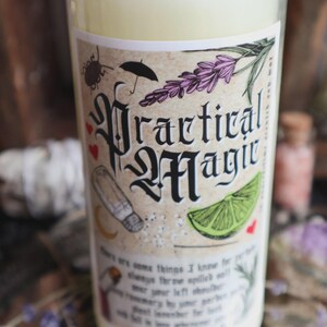 Peut inclure: Une bougie cylindrique en cire couleur crème avec une étiquette portant le texte Practical Magic en lettres gothiques noires. L'étiquette présente des illustrations d'une salière, d'un quartier de citron vert, de lavande, d'un parapluie et de cœurs rouges.