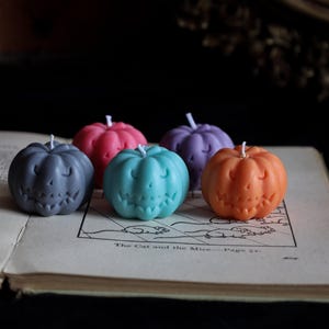 May include: Five small pumpkin-shaped candles in grey, pink, teal, purple, and orange. Each candle features a carved jack-o'-lantern face with a smiling expression and a single white wick at the top, arranged in a cluster on an open vintage book.