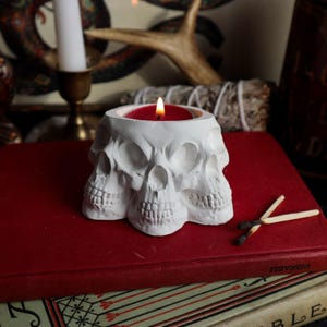 May include: A white matte skull-shaped candle holder featuring three conjoined craniums arranged in a circular formation. A small red tea light candle is lit inside the central cavity, emitting a warm flame. The object is crafted from a solid material.