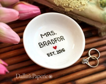 Puede incluir: Plato de cerámica blanco para anillos con letras negras que dicen "MRS. BRADFOR EST. 2016" con un corazón rojo. Dos anillos de oro están en el plato.