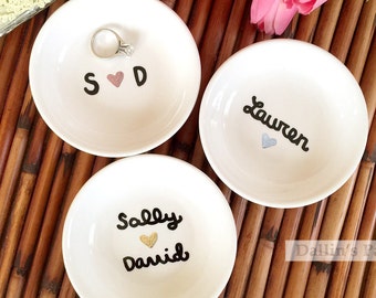 Puede incluir: Tres platos de anillos de cerámica blanca con letras negras. El primer plato dice "S♡D" con un corazón rosa. El segundo plato dice "Lauren" con un corazón plateado. El tercer plato dice "Sally♡David" con un corazón dorado.