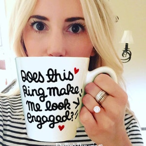 May include: White ceramic mug with black lettering that reads "Does this ring make me look engaged?" with two red hearts. A person is holding the mug and wearing a silver ring with a large clear stone.