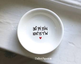 Puede incluir: Plato de anillo de cerámica blanca con coordenadas escritas a mano en negro "30° 39' 33"N 104° 15' 7"W" y un pequeño corazón rojo.