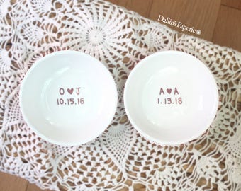 Puede incluir: Dos cuencos de cerámica blanca con letras rosas. Los cuencos están personalizados con iniciales y fechas. El primer cuenco dice "O ♥ J 10.15.16" y el segundo cuenco dice "A ♥ A 1.13.18".