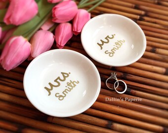 Puede incluir: Dos platos de anillos de cerámica blanca con letras doradas. Los platos dicen "Mrs. Smith" y "Mr. Smith". Un par de anillos de plata están sobre una superficie de madera.