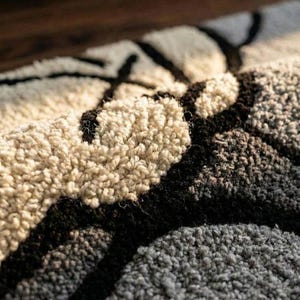 May include: Close-up of a textured rug with a geometric pattern. The rug features a combination of beige, black, and gray colors. The design appears to be abstract, with thick, dark lines contrasting against the lighter background.