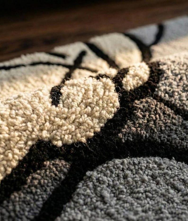 May include: Close-up of a textured rug with a geometric pattern. The rug features a combination of beige, black, and gray colors. The design appears to be abstract, with thick, dark lines contrasting against the lighter background.