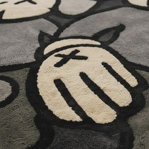 May include: A tufted area rug featuring a repeating graphic pattern of cartoon-style gloved hands and circular faces with X-shaped eyes. The design is rendered in a monochromatic palette of cream, light gray, dark gray, and bold black outlines.