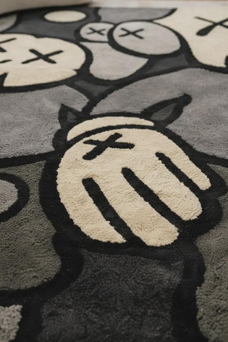 May include: A tufted area rug featuring a repeating graphic pattern of cartoon-style gloved hands and circular faces with X-shaped eyes. The design is rendered in a monochromatic palette of cream, light gray, dark gray, and bold black outlines.