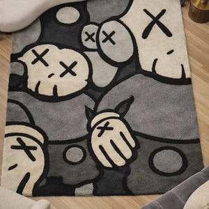 May include: A rectangular area rug featuring a graphic pattern of stylized cartoon characters in shades of gray, black, and cream. The design includes multiple faces with X-shaped eyes and gloved hands with X symbols, rendered in a thick, textured pile.