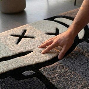 May include: A modern, abstract rug featuring a face design. The rug has a white face with black cross-shaped eyes and a black mouth, set against a gray background. The rug's texture appears soft and plush.