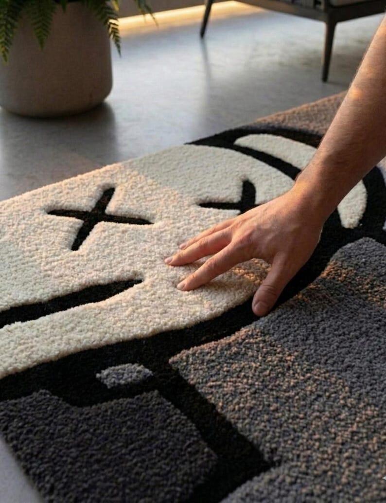 May include: A modern, abstract rug featuring a face design. The rug has a white face with black cross-shaped eyes and a black mouth, set against a gray background. The rug's texture appears soft and plush.