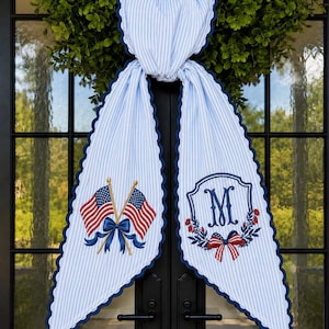 May include: A light blue and white pinstriped fabric door hanger with scalloped navy blue edges. One side features an embroidered pair of crossed American flags with a navy bow, while the other side displays a navy letter M inside a floral wreath.