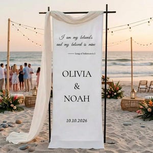 May include: A white banner with the text "I am my beloved's and my beloved is mine - Song of Solomon 6:3". Below, the names "OLIVIA & NOAH" are displayed, followed by the date "10.10.2026". The banner is part of a beach wedding setup.