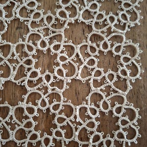 Può includere: Primo piano di un centrino in pizzo fatto a mano color crema. L'intricato design presenta un motivo ripetuto di anelli e curve, creando una superficie delicata e strutturata. Il centrino è posto su una superficie di legno.