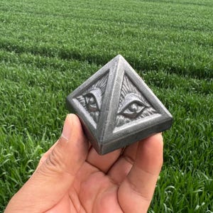 Può includere: Una scultura a forma di piramide grigio scuro con due occhi stilizzati all'interno di un triangolo. La scultura ha una superficie strutturata ed è tenuta contro uno sfondo di erba verde. La piramide è un oggetto decorativo unico.