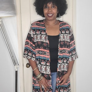 Op de afbeelding: Een vrouw draagt een lange, zwart-witte kimono met rode en blauwe accenten. De kimono heeft een V-hals en is open aan de voorkant. Ze draagt een zwart tanktop en een blauwe jeans.