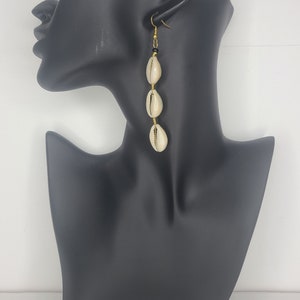 May include: Dangling earrings featuring three cream-colored cowrie shells, strung together with gold-colored wire and a small black bead. The earrings hang from a gold-colored hook. The earrings are displayed on a black mannequin.