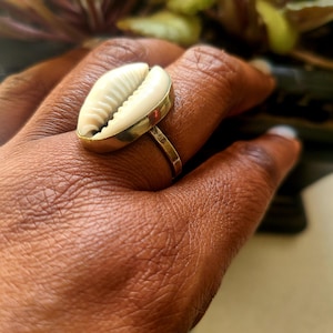 May include: A gold ring with a white seashell design. The ring is worn on a person's finger.
