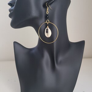 May include: Gold-tone hoop earrings with a natural seashell pendant. The earrings feature black beads and a gold-tone hook closure. The seashell is wrapped with gold wire. The earrings are displayed on a black mannequin head.