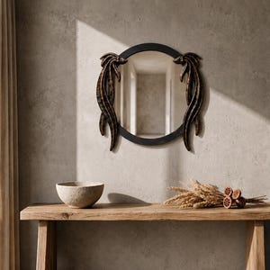 Puede incluir: Un espejo de pared redondo con un marco oscuro y texturizado, que presenta dos elementos decorativos simétricos y alargados en tonos bronce a los lados. El espejo tiene una superficie reflectante lisa y un borde negro circular.